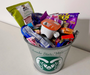 A tin bucket with the Rams head logo with the words "Colorado State University" on it. Inside, the bucket is filled with snacks, like Pringles, Takis, candy, peanuts and more.