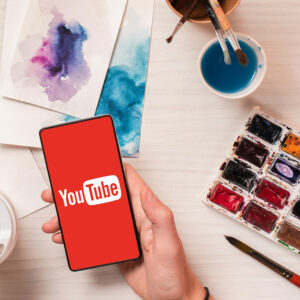 A hand holds a phone with a red background and YouTube logo. Surrounding the hand are painting tools and drawings.
