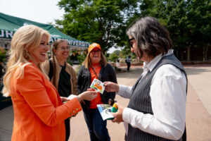 ALT TEXT said: President Amy Parsons and Dean Lawrence Dill smile and exchange C S U-themed stickers during a sticker drop event on campus. Two other people stand nearby, also smiling. A green tent with partially visible "Colorado State University" text is in the background, along with trees and a campus walkway.